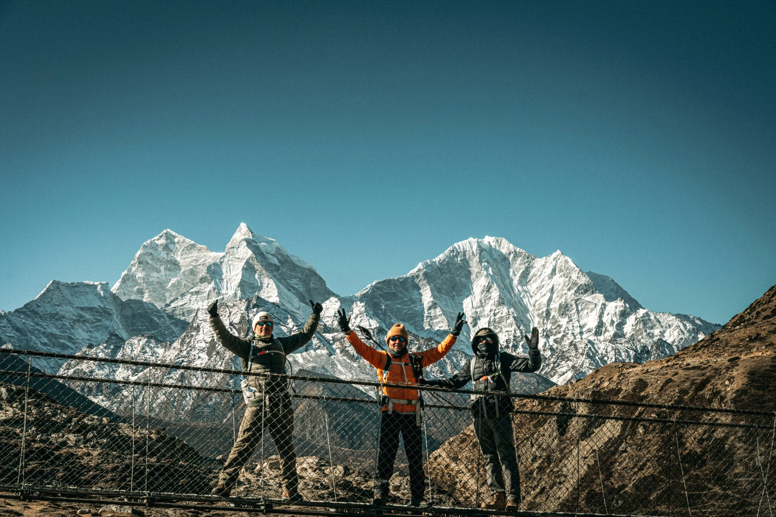 Three trekkers on a suspension bridge with their arms raised, Himalayan peaks behind them