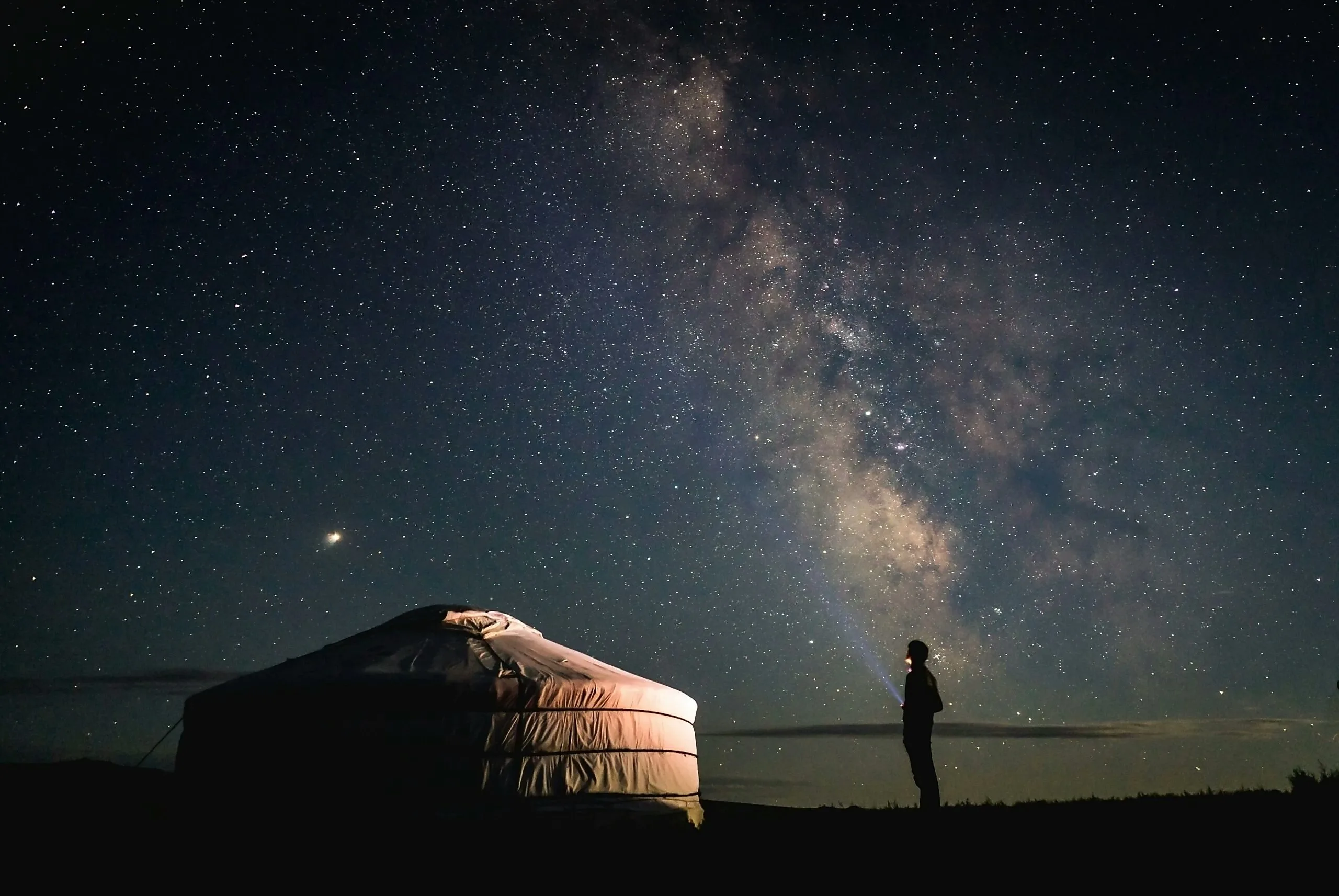 A traveller under the Milky Way beside a lit yurt at night