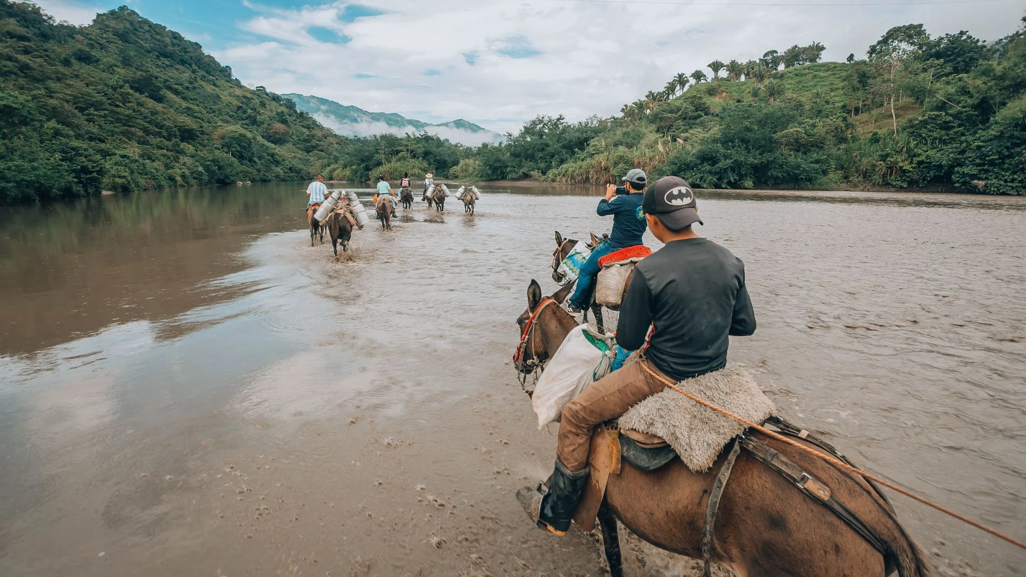 Riders fording a wide river below forested hills