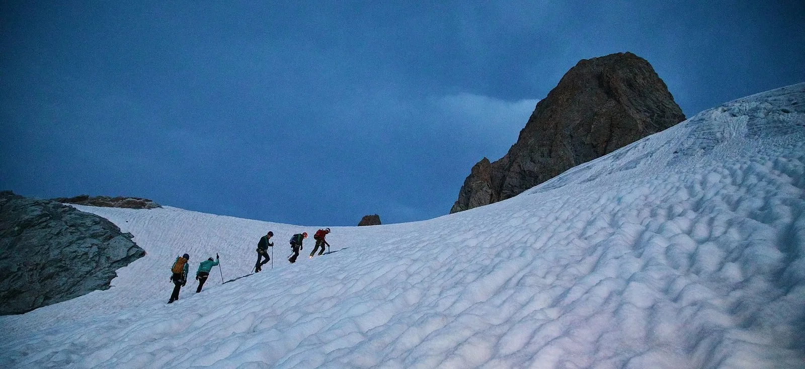 Five climbers roped together crossing a snowfield at dusk, a rock spire above them