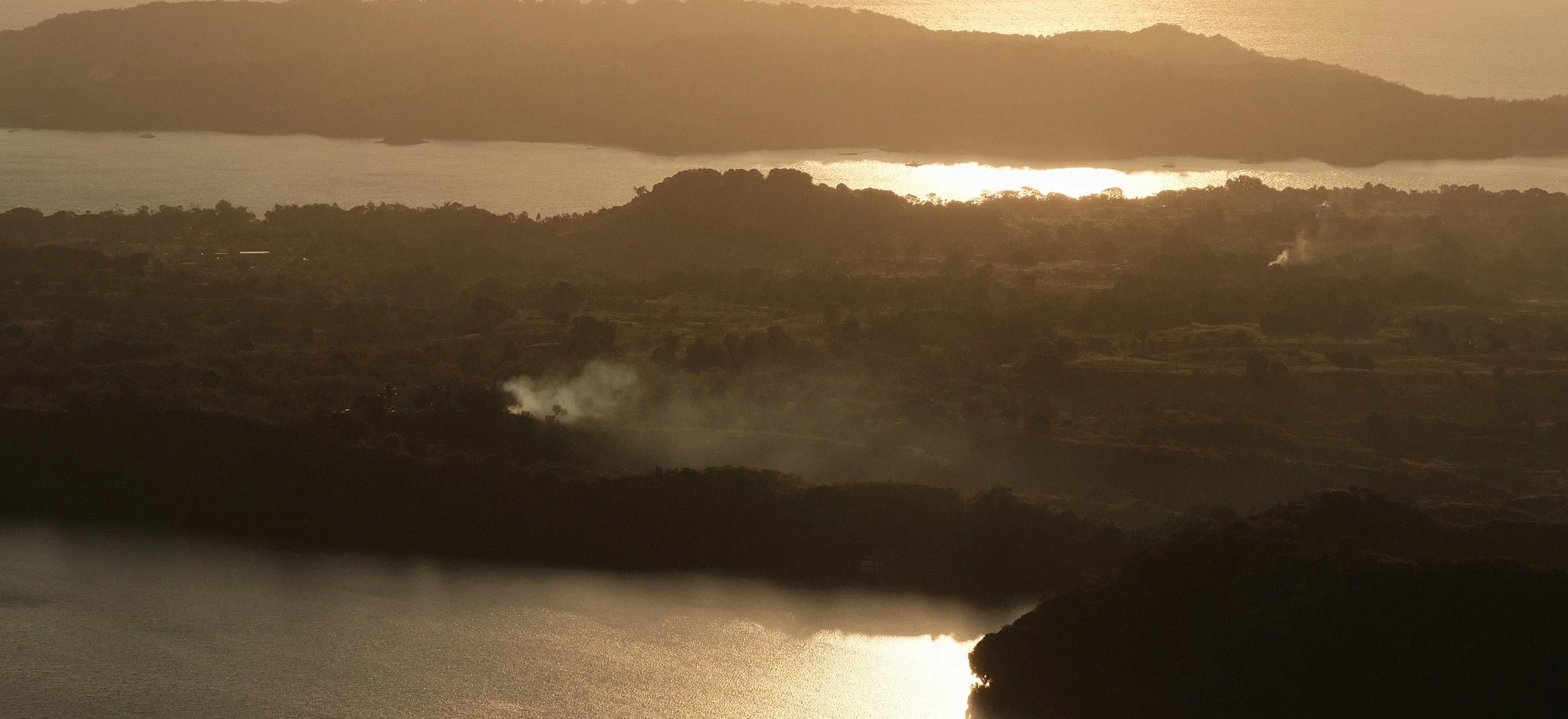 Forested hills above an inlet in hazy golden light, smoke rising from the trees