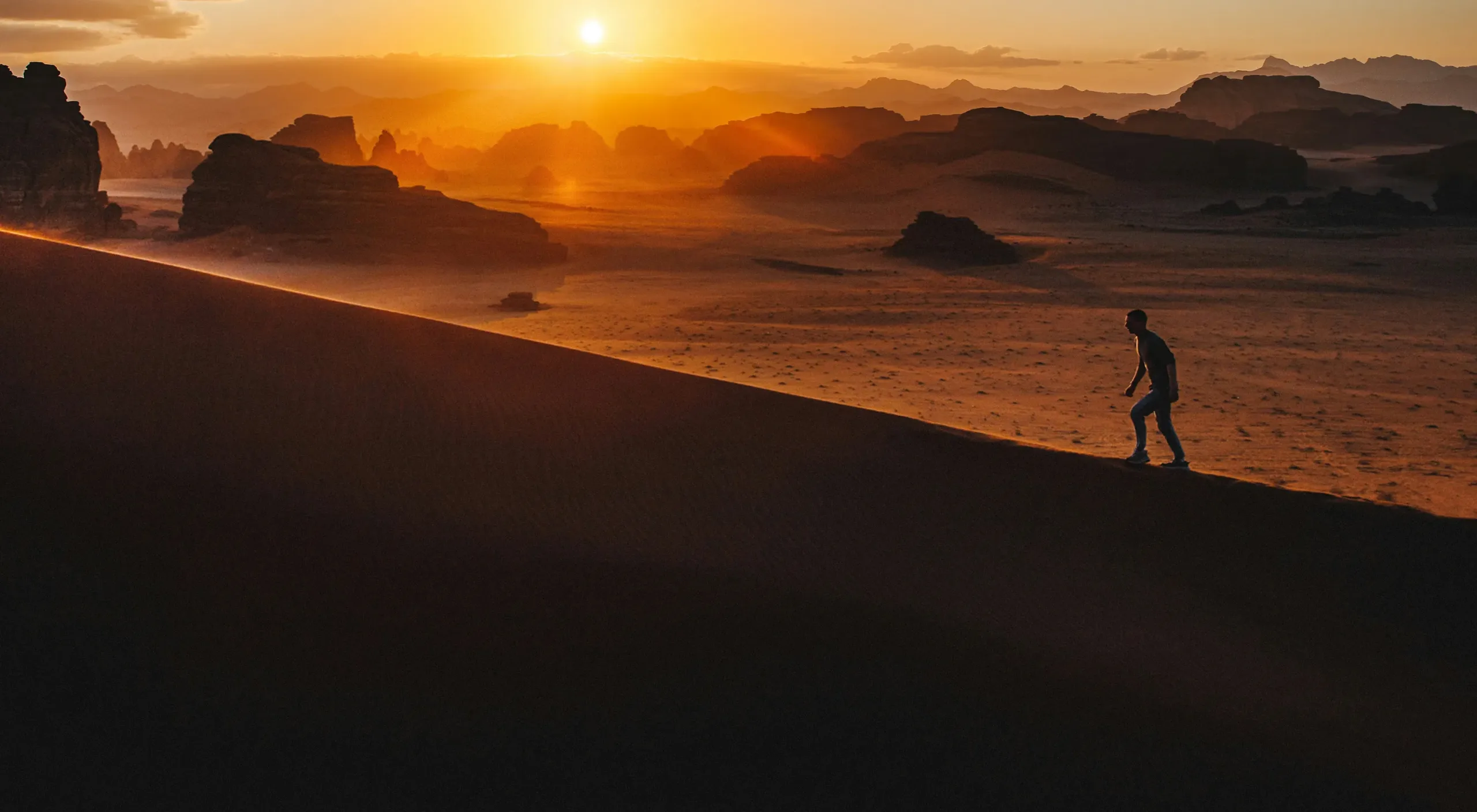 A walker climbing a dune ridge at sunset, with desert rock towers behind